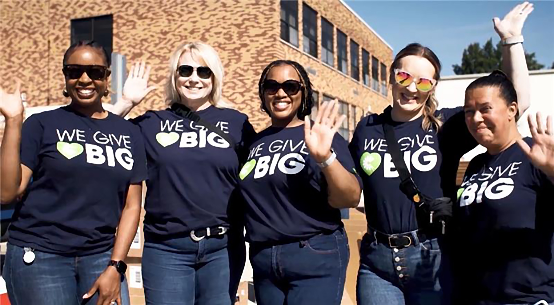 Group of volunteers wearing “We Give Big” shirts smile and wave during a community service event in Michigan.